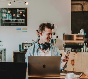 Young man smiling at his phone while sitting at coffee shop with laptop